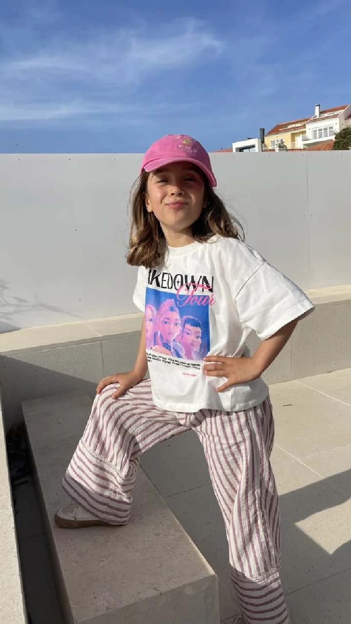 Full-body photo of a girl in a pink cap, white printed t-shirt and striped trousers